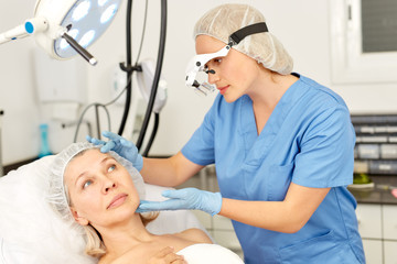 Professional cosmetician examining face skin of mature woman in clinic of esthetic cosmetology