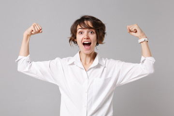 Excited strong young business woman in white shirt posing isolated on grey wall background studio portrait. Achievement career wealth business concept. Mock up copy space. Showing biceps, muscles.