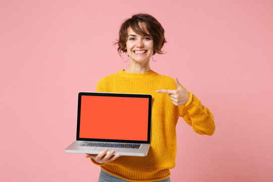 Smiling young brunette woman girl in yellow sweater posing isolated on pink background. People lifestyle concept. Mock up copy space. Point index finger on laptop pc computer with blank empty screen.