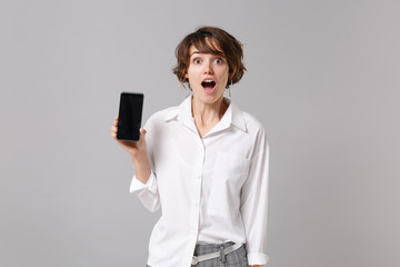 Naklejka premium Shocked young business woman in white shirt posing isolated on grey background in studio. Achievement career wealth business concept. Mock up copy space. Holding mobile phone with blank empty screen.