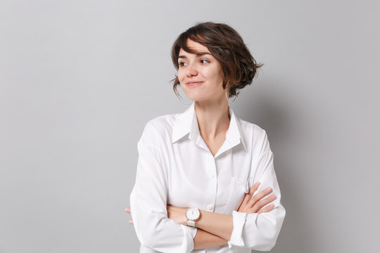 Smiling young business woman in white shirt posing isolated on grey background studio portrait. Achievement career wealth business concept. Mock up copy space. Holding hands crossed, looking aside.