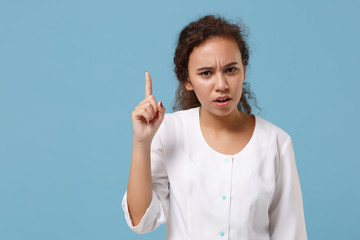 Concerned african american doctor woman isolated on blue wall background. Female doctor in white medical gown holding index finger up. Healthcare personnel health medicine concept. Mock up copy space.