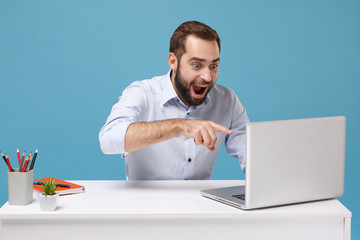 Excited young bearded man in light shirt sit at desk isolated on pastel blue background. Achievement business career concept. Mock up copy space. Work on project, point finger on laptop pc computer.