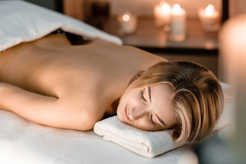 Obraz na płótnie Canvas Spa concept. Young beautiful slim girl lies on massage table in anticipation of massage on a background of blurry burning candles.Front view, copy space