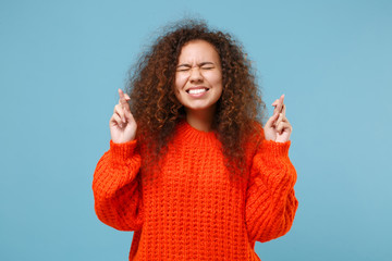 Young african american girl in casual orange knitted clothes keep fingers crossed eyes closed...