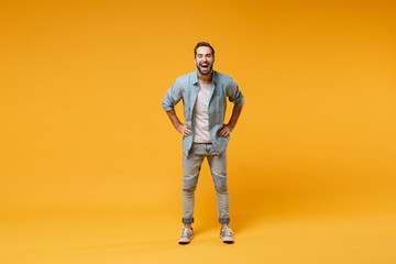 Laughing young bearded man in casual blue shirt posing isolated on yellow orange background, studio portrait. People emotions lifestyle concept. Mock up copy space. Standing with arms akimbo on waist.