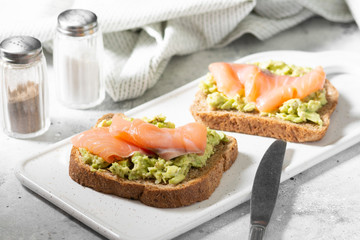 Bruschetta with avocado and salmon on a white ceramic plate on a light gray kitchen table. Toasted bread with avocado and red fish. The concept is helpful and a delicious Breakfast