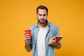 Perplexed displeased young student man in casual blue shirt posing isolated on yellow orange background in studio. People lifestyle concept. Mock up copy space. Holding notebook, cup of coffee or tea.