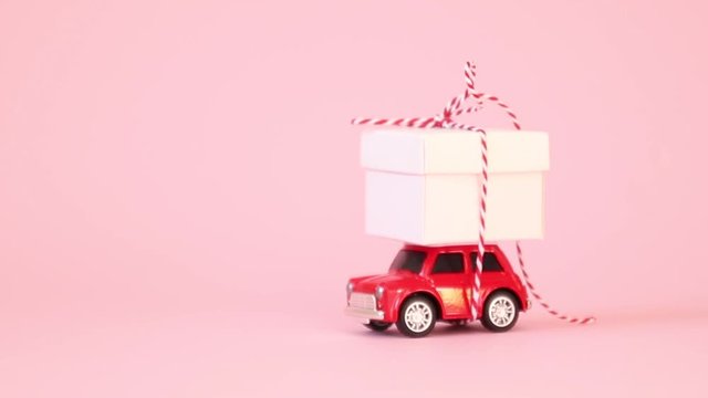 Red retro toy car delivery gift box with ribbon bow on a pink background. Colorful confetti falls on a car. Birthday, Valentine's Day, Women's Day concept.
