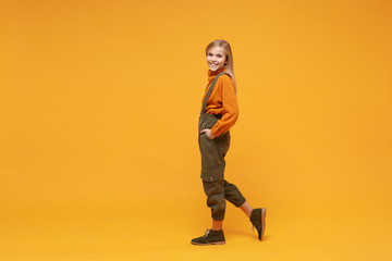 Side view of smiling little blonde kid girl 12-13 years old in turtleneck jumpsuit isolated on orange yellow wall background in studio. Childhood lifestyle concept. Mock up copy space. Looking camera.