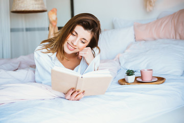 Fototapeta na wymiar Girl reading a book, drinking coffee or tea, resting in bed.
