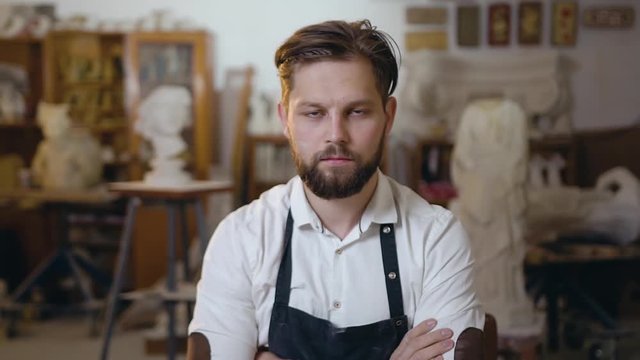 Handsome 30-aged serious bearded stonemason dressed in workwear sitting at his workplace in studio and posing on camera