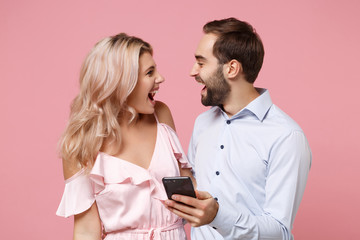 Cheerful young couple two guy girl in party outfit celebrating isolated on pastel pink background. Valentine's Day Women's Day birthday holiday party concept. Using mobile phone, typing sms message.