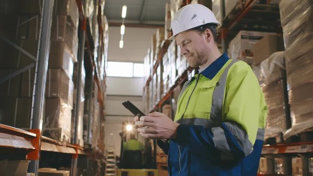 Side view of warehouse worker surfing internet on smartphone standing in warehouse