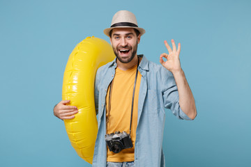 Funny traveler tourist man in yellow clothes with photo camera isolated on blue background. Passenger traveling abroad on weekends. Air flight journey concept. Hold inflatable ring showing OK gesture.