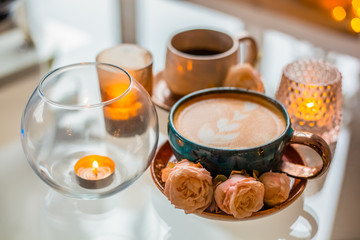 A cup of coffee on a saucer is decorated with roses next to candles in beautiful candlesticks on a glass table