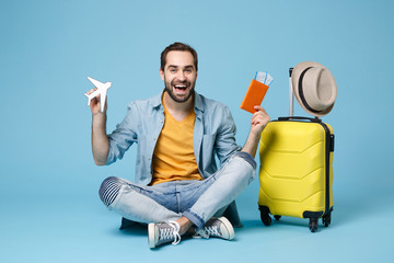 Cheerful traveler tourist man in yellow clothes isolated on blue background. Passenger traveling abroad on weekend. Air flight journey Sit near suitcase hold passport boarding pass tickets air plane.