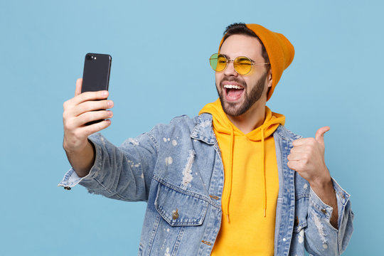 Funny Young Hipster Guy In Fashion Jeans Denim Clothes Isolated On Pastel Blue Background. People Lifestyle Concept. Mock Up Copy Space. Doing Selfie Shot On Mobile Phone, Showing Thumb Up, Blinking.