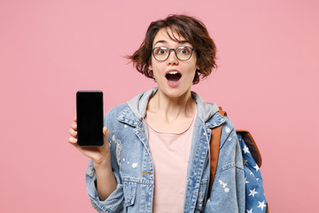 Surprised young woman student in denim clothes, eyeglasses, backpack isolated on pastel pink background. Education in high school university college concept. Hold mobile phone with blank empty screen.