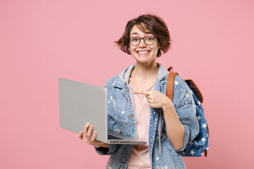 Fototapeta Smiling young woman student in denim clothes eyeglasses backpack isolated on pastel pink background. Education in high school university college concept. Pointing index finger on laptop pc computer. obraz