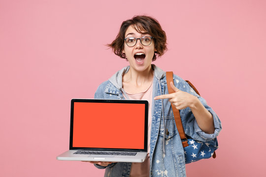 Excited girl student in denim clothes glasses backpack isolated on pink background. Education in high school university college concept. Point index finger on laptop computer with blank empty screen.