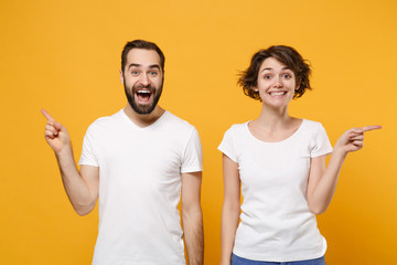 Cheerful young couple friends bearded guy girl in white blank empty t-shirts posing isolated on yellow orange background. People lifestyle concept. Mock up copy space. Pointing index fingers aside up.