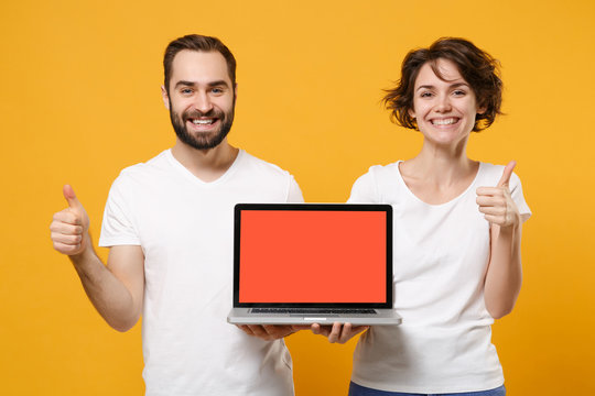 Smiling couple friends bearded guy girl in white t-shirts isolated on yellow orange background. People lifestyle concept. Mock up copy space. Hold laptop computer with blank screen, showing thumb up.