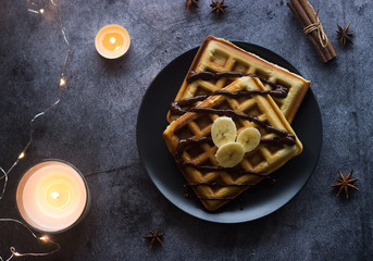 Viennese waffles on a plate with banana and chocolate. In the background are lights and candles. Romantic picture