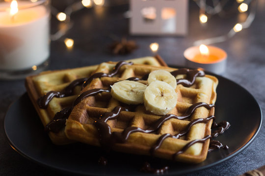 Viennese waffles on a plate with banana and chocolate. In the background are lights and candles. Romantic picture