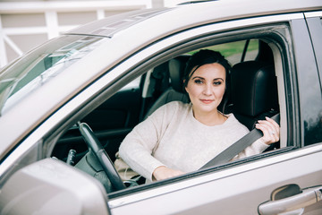 dark haired brunette woman sitting in the driver's seat of a modern tan sedan car and putting on her seat belt