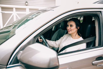 dark haired brunette woman sitting in the driver's seat of a modern tan sedan car and putting on...