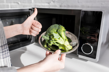 cropped view of woman holding plate with broccoli near microwave and showing like