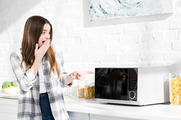 shocked and attractive woman pointing with finger at microwave in kitchen
