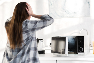 Fototapeta na wymiar back view of shocked woman looking at broken microwave in kitchen