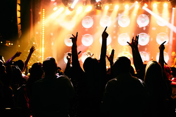 Crowd at a music concert, audience raising hands up in front of bright stage lights.