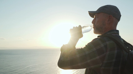 senior traveler drinking pure water from bottle in front of sunrise over a sea