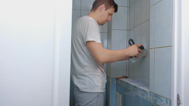 Man tiler making renovation repair at home, he removes tiles from wall in toilet using tool spatula. Construction works at home. He removes seams between tiles to remove it.