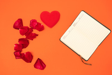 Notebook on orange background, red rose petal and red heart on orange background,