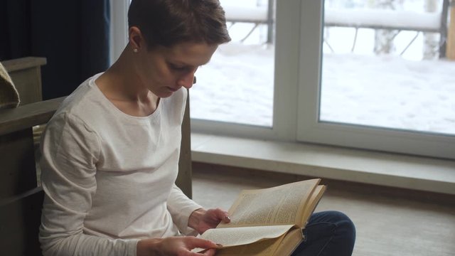 A young girl with short hair is reading a book while sitting on the floor at home by the window on a frosty winter evening. Woman enjoying literature. Female education and studying. Holidays in chalet