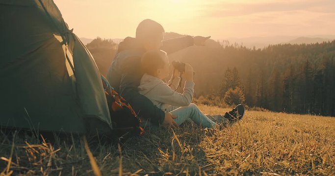 Happy traveler father and son sitting on grass and look through binoculars, bonding around mountain under sun light enjoying the leisure and freedom. 4K slow motion video