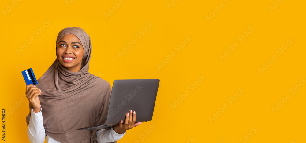 Wall mural doubtful afro muslim girl holding credit card and laptop in hands