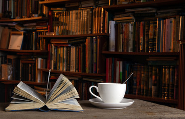 Open book. Nearby is a cup of coffee on a saucer and with a teaspoon. In the background shelves...
