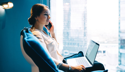 Thoughtful pleased businesswoman phoning while interacting with laptop in light contemporary office