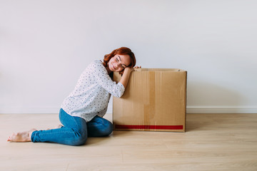 Attractive girl moving into the new apartment. Sitting on the floor with carton box in an empty room, smiling. Daydreaming, white wall background.