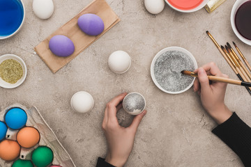 cropped view of woman decorating chicken eggs with silver glitter on grey concrete surface