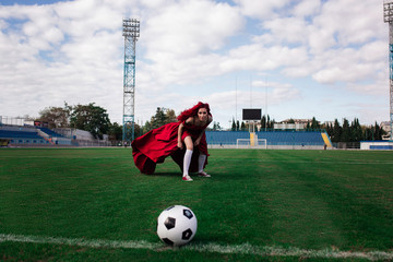 Luxurious woman with red hair and in a red dress plays on the football field.