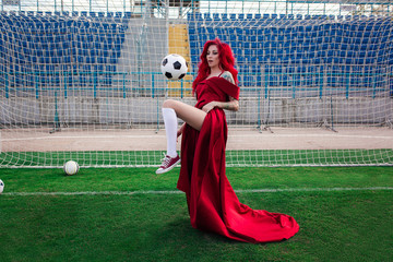 Luxurious woman with red hair and in a red dress plays on the football field.