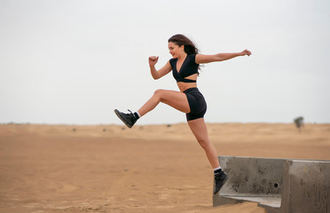 Fototapeta premium Beautiful long hair latin brunette fitness model wearing black short fitness outfit exercising and leaping in the middle eastern desert sand