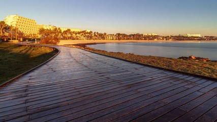 Wooden Boardwalk at sunrise leading to holiday resort lit by morning sun, beach and bay with hotels, restaurants, cafes and palm trees, Sa Coma, Mallorca, Spain.