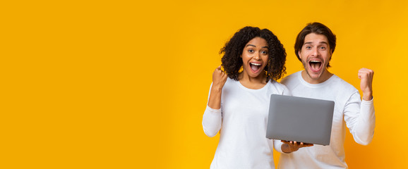 Cheerful multiracial couple celebrating success with laptop, raising fists and shouting